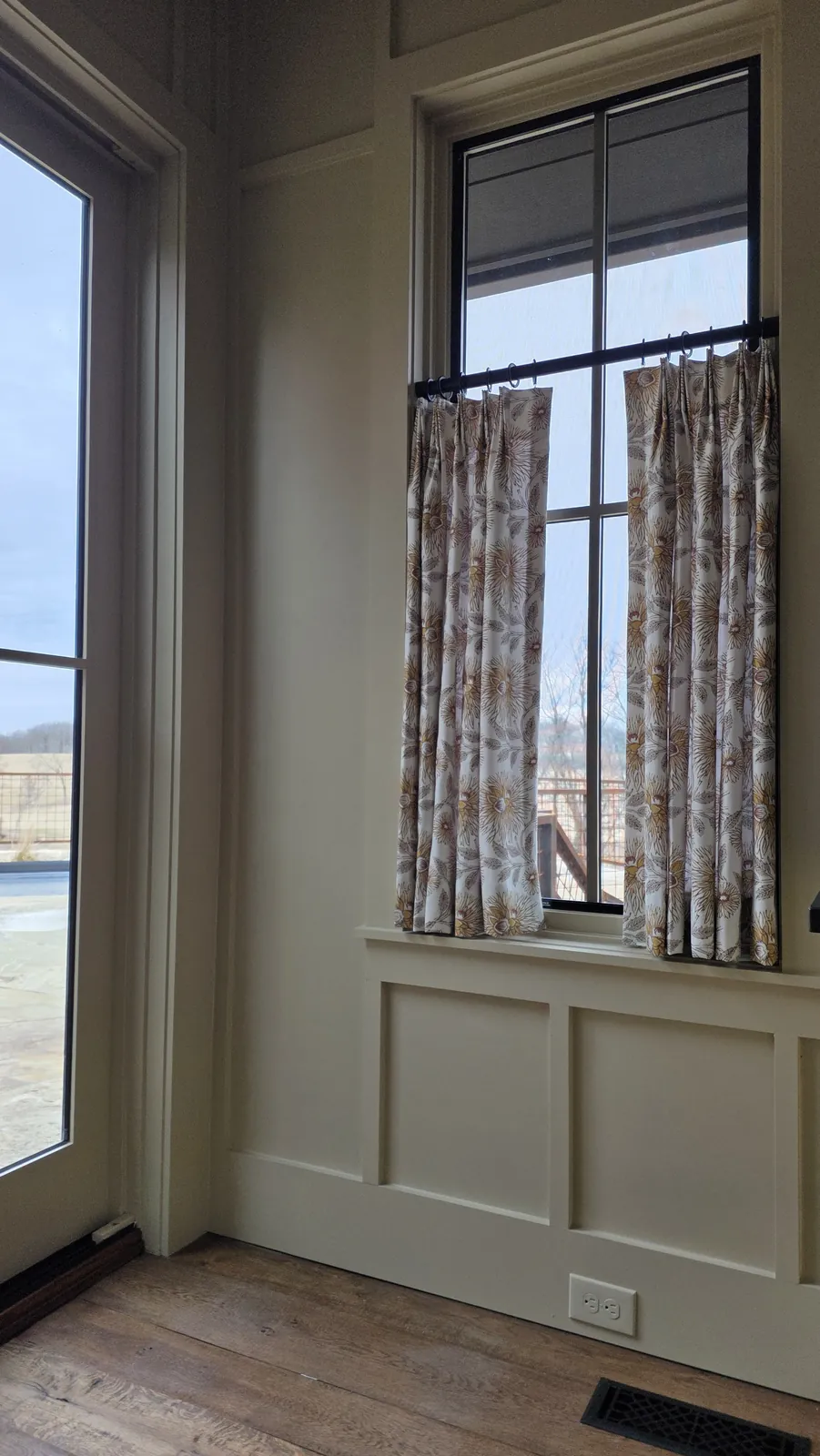 Café curtains on a tall window above a paneled wall, installed by Solar Shield Blinds