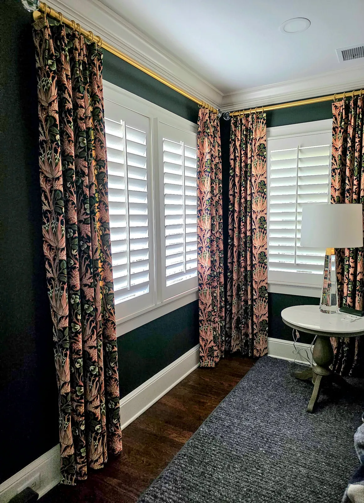 Floral print drapery panels over plantation shutters in a dark green room, installed by Solar Shield Blinds