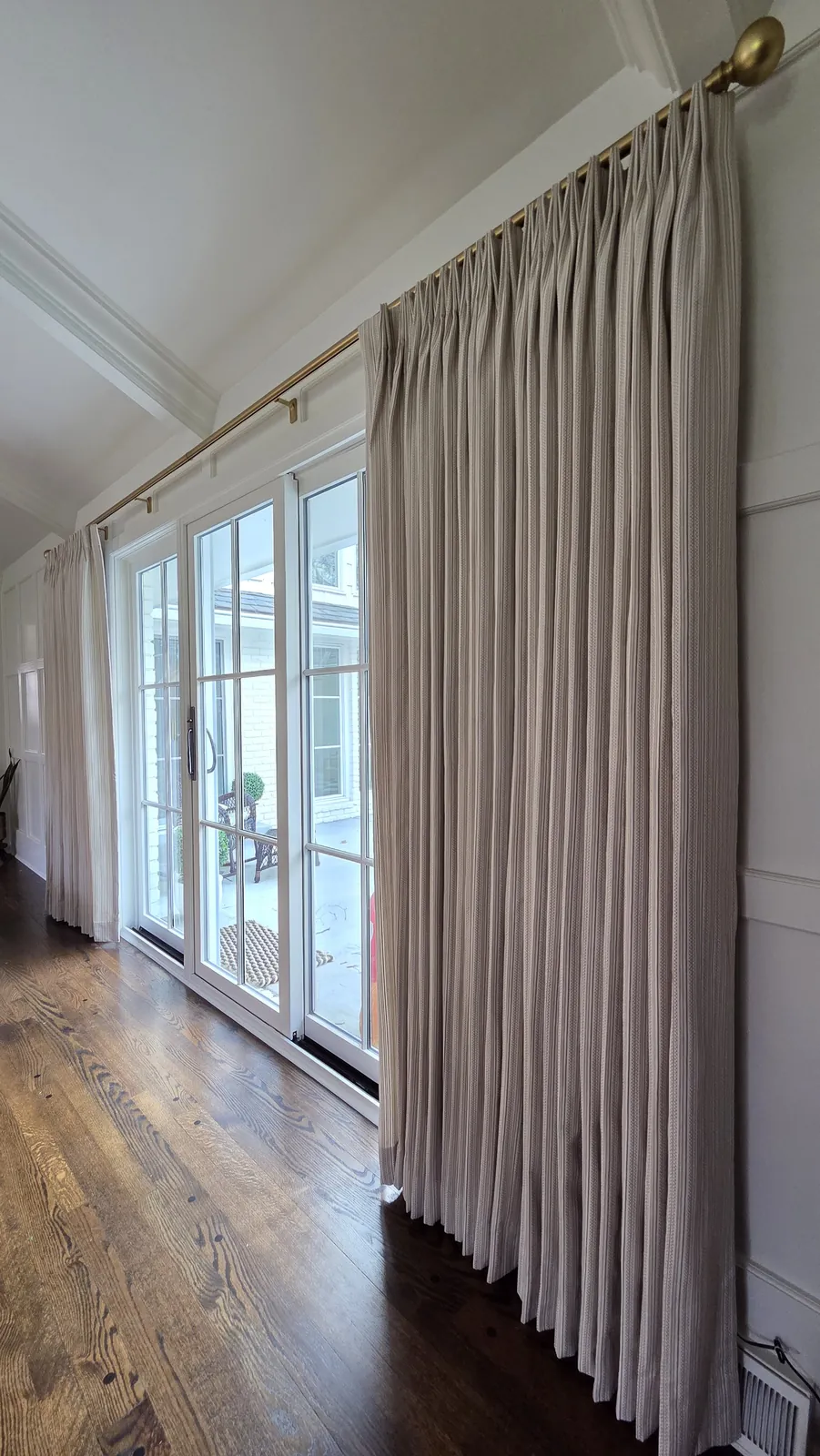 Pinch pleat drapery panel on a brass rod beside French doors, installed by Solar Shield Blinds