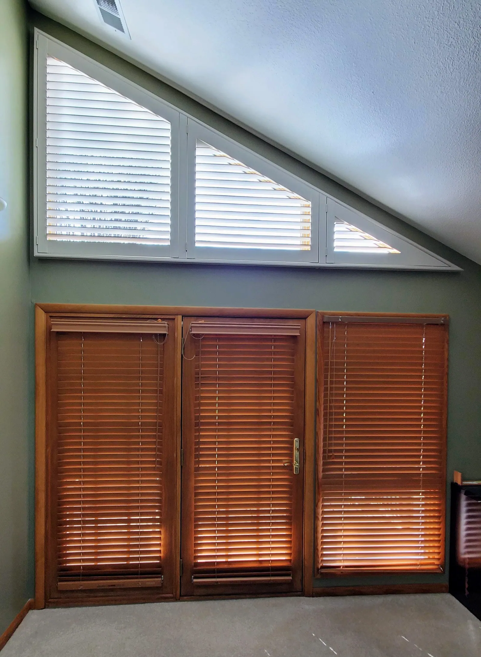 Plantation shutters built to a sloped ceiling window, above wood blinds on a set of doors