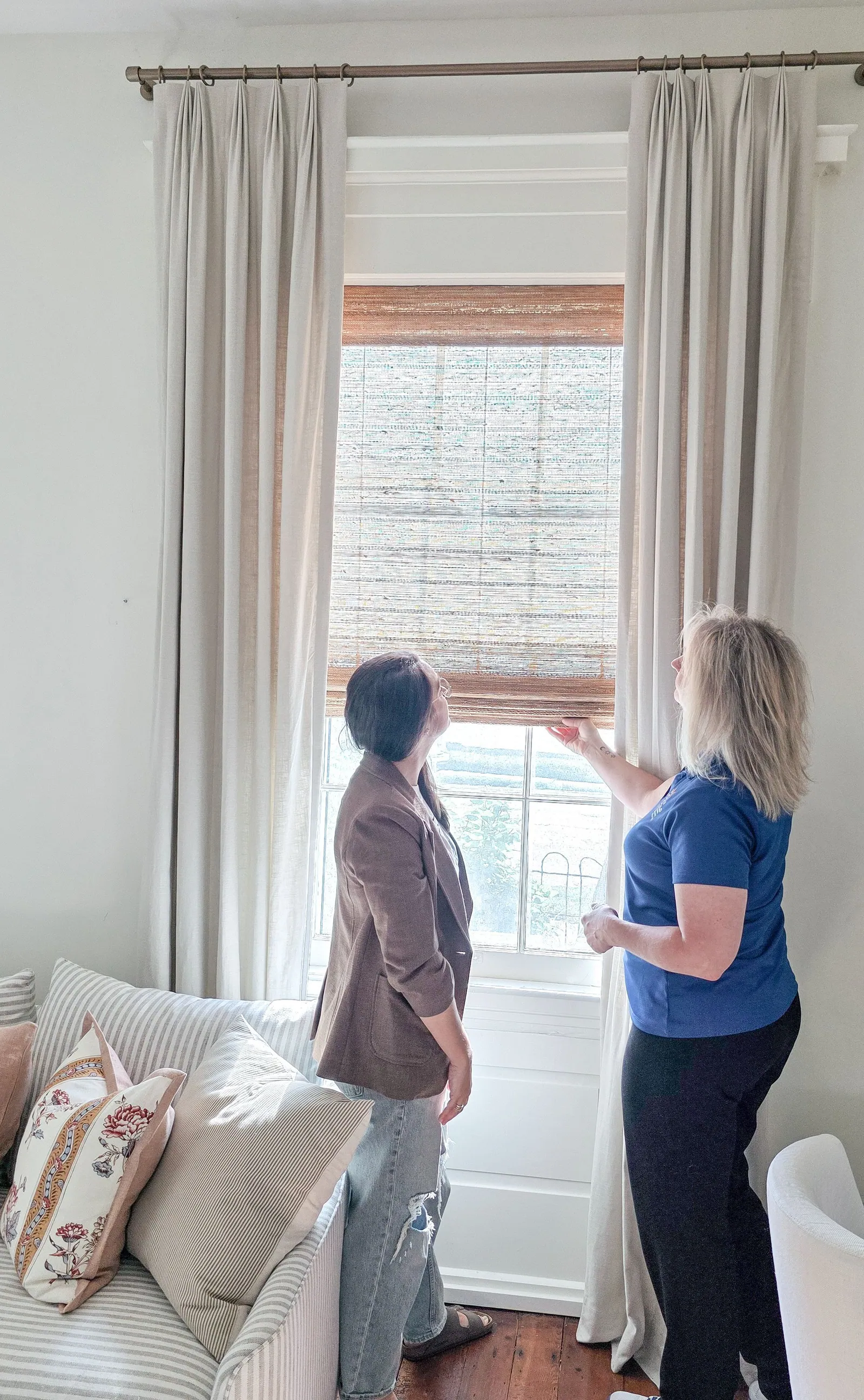 A designer and a Solar Shield Blinds specialist adjusting a woven shade behind drapery panels