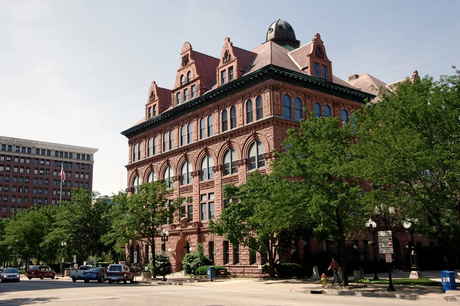 A red brick civic building with towers and arched windows, framed by trees