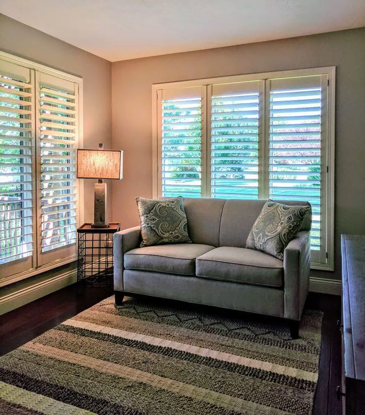 White plantation shutters on two windows behind a gray loveseat