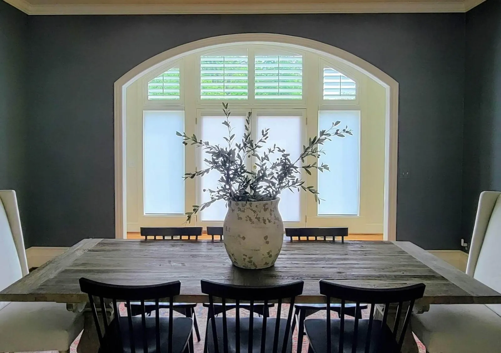 Plantation shutters in an arched transom over French doors fitted with shades, seen across a dining table