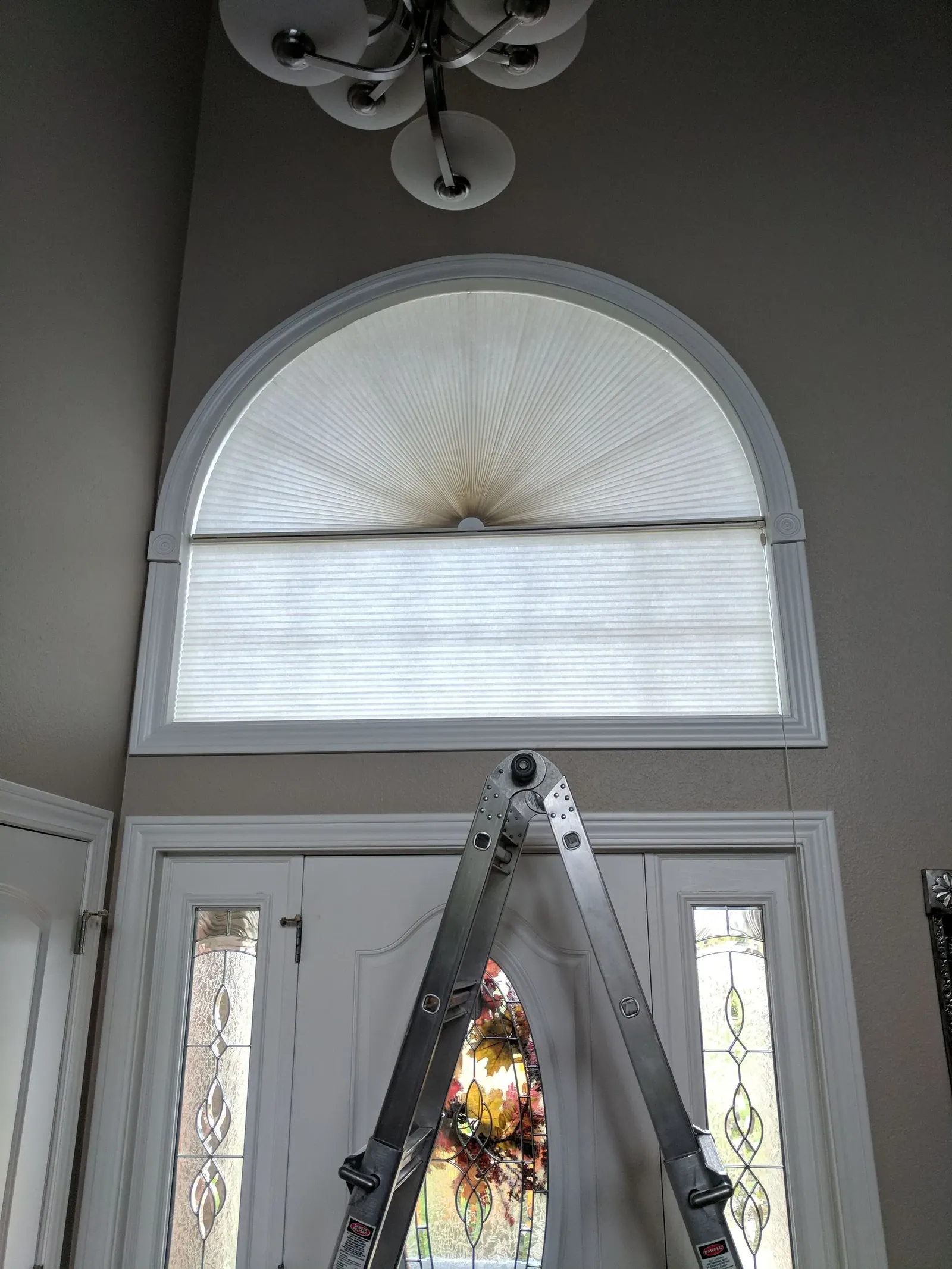A ladder set under the half round window above a front door, with a pleated fan shade fitted in the arch
