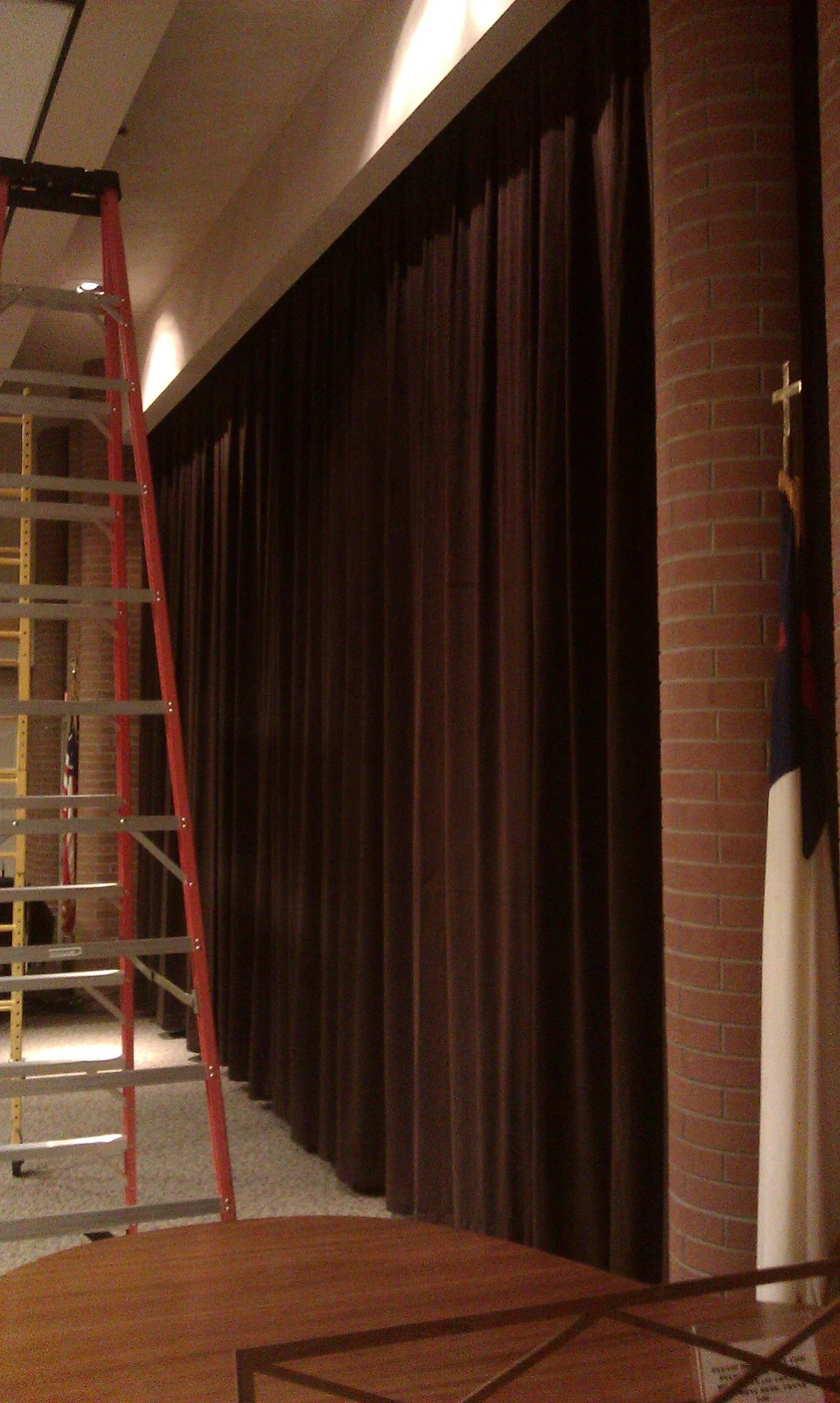 Floor to ceiling brown pleated drapes along a brick wall in a community room, with a ladder set up for the install