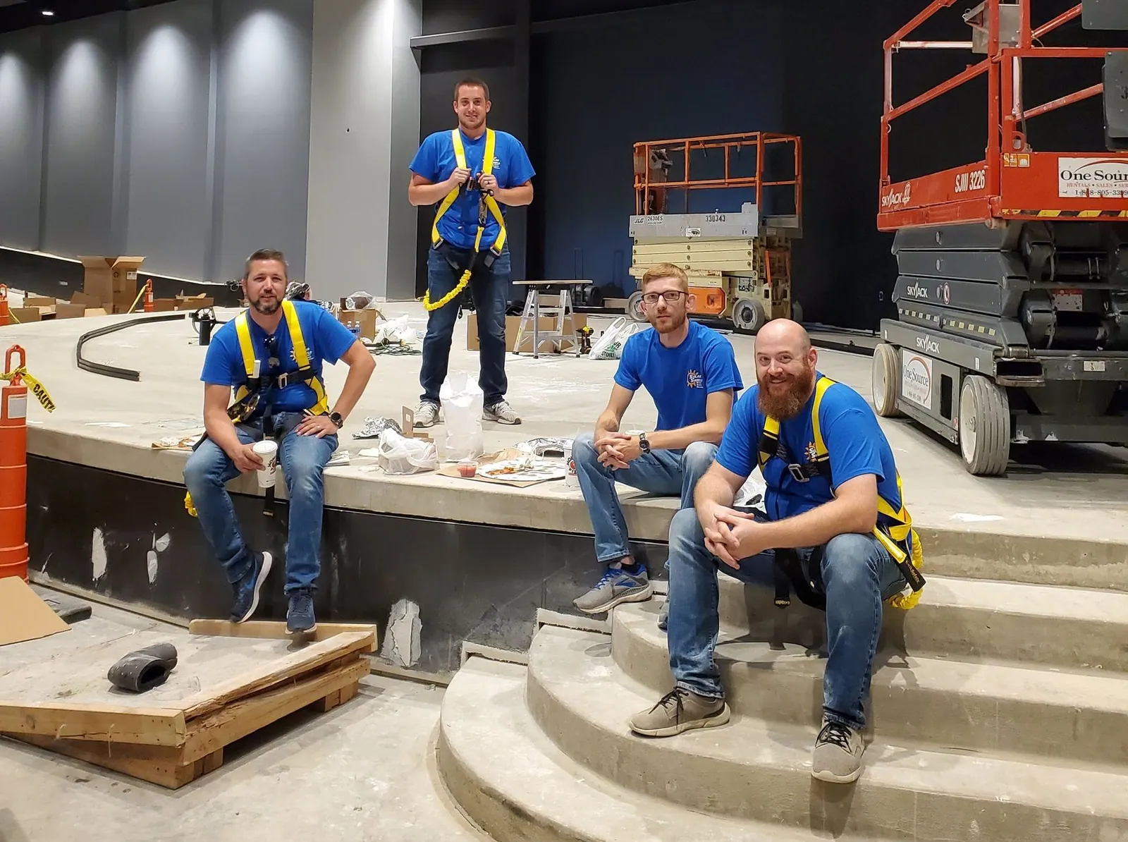 Four Install Techs in blue Solar Shield Blinds shirts and safety harnesses on the edge of a stage under construction, with a scissor lift behind them