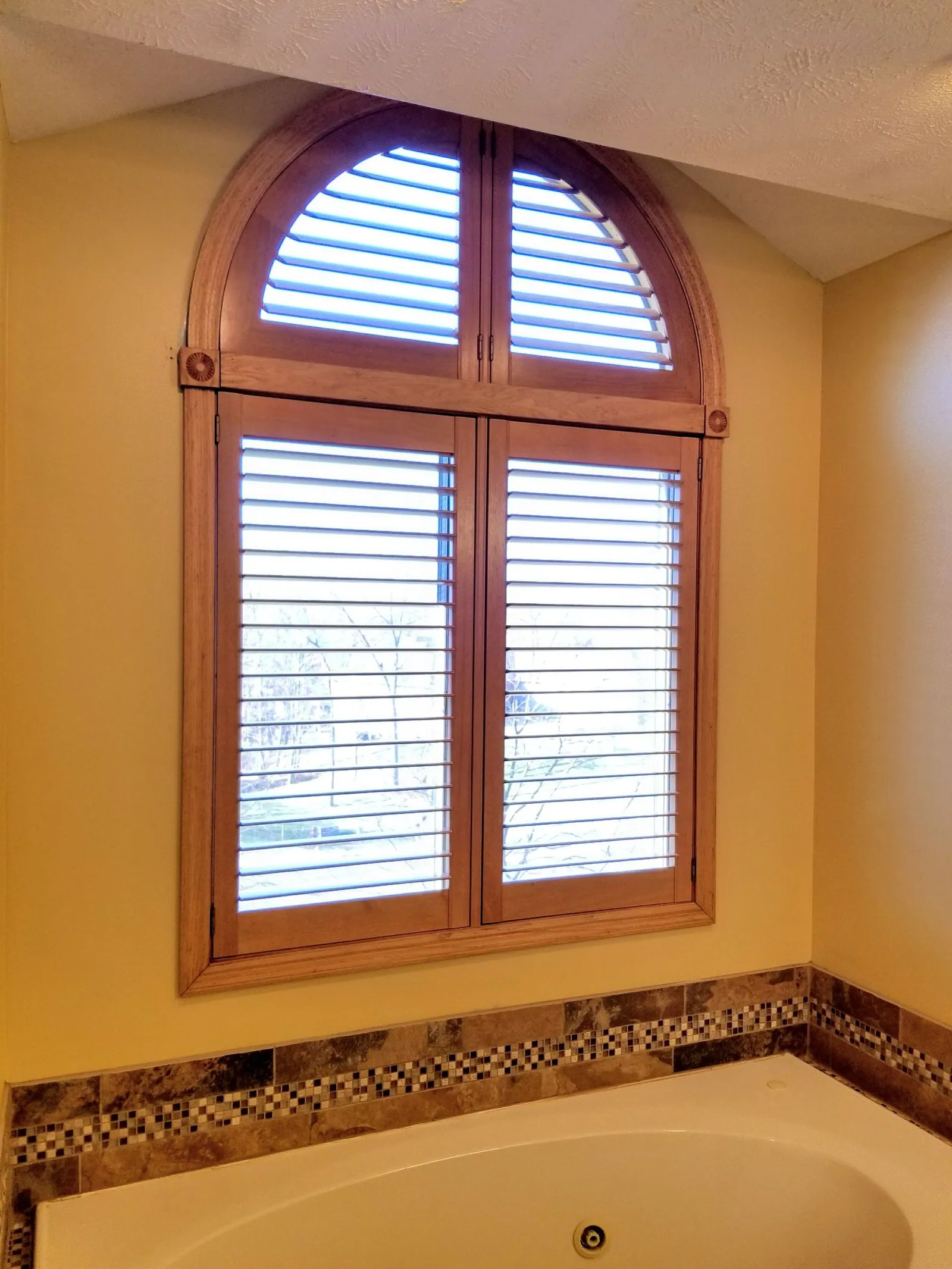 Stained wood arch top plantation shutter over a tiled bathtub
