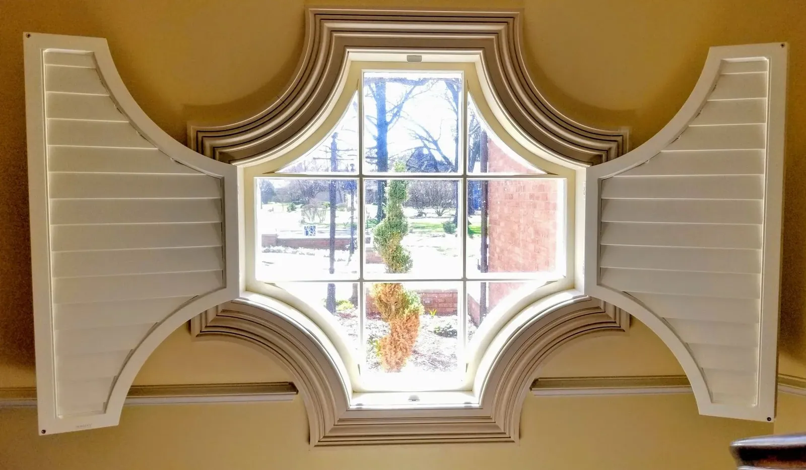 Scalloped plantation shutter seen from inside with the louvers open to the trees